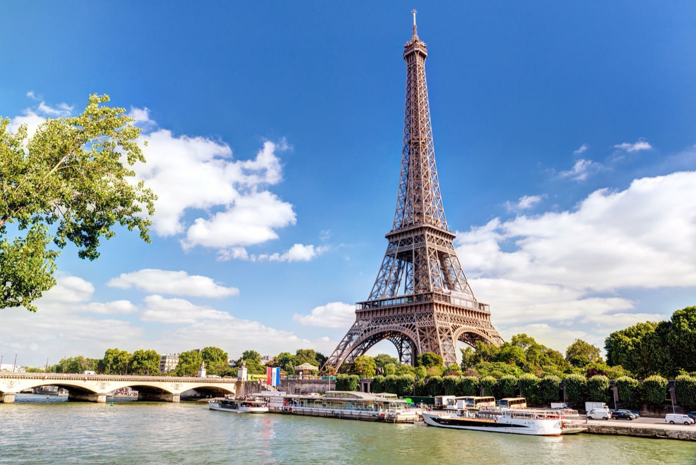 Der Eiffelturm im Sommer von der Seine aus fotografiert, mit Ausflugsbooten auf dem Wasser und einer Brücke