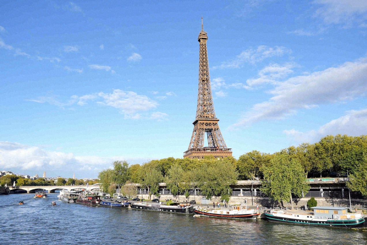 Blick auf den Eiffelturm von der Seine aus, mit Hausbooten und einer Brücke im Vordergrund.